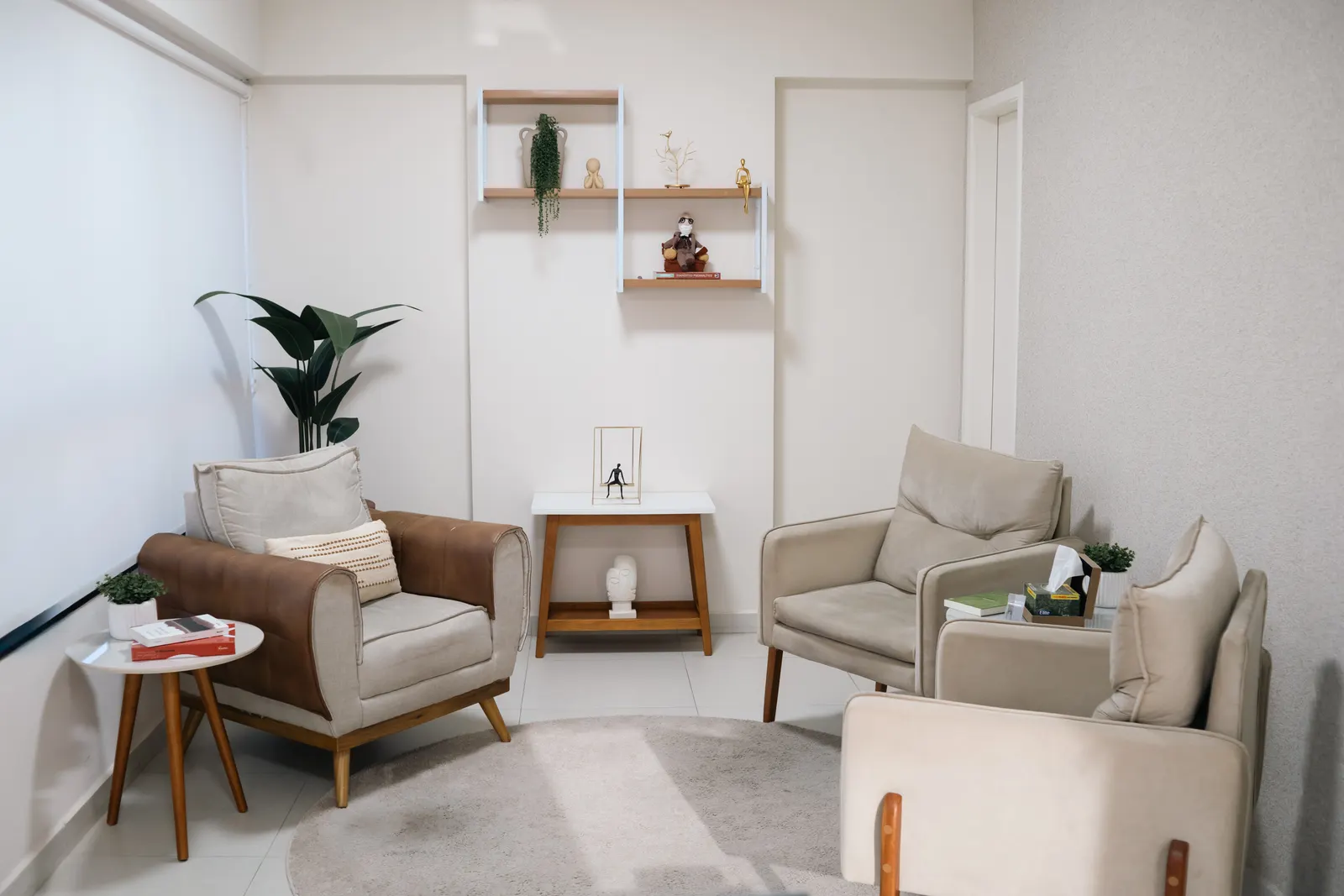 Sala de atendimento com poltronas de couro e tecido bege dispostas ao redor de um tapete redondo, prateleiras de madeira com objetos decorativos na parede e uma caixa de lenços sobre a mesinha entre as cadeiras.