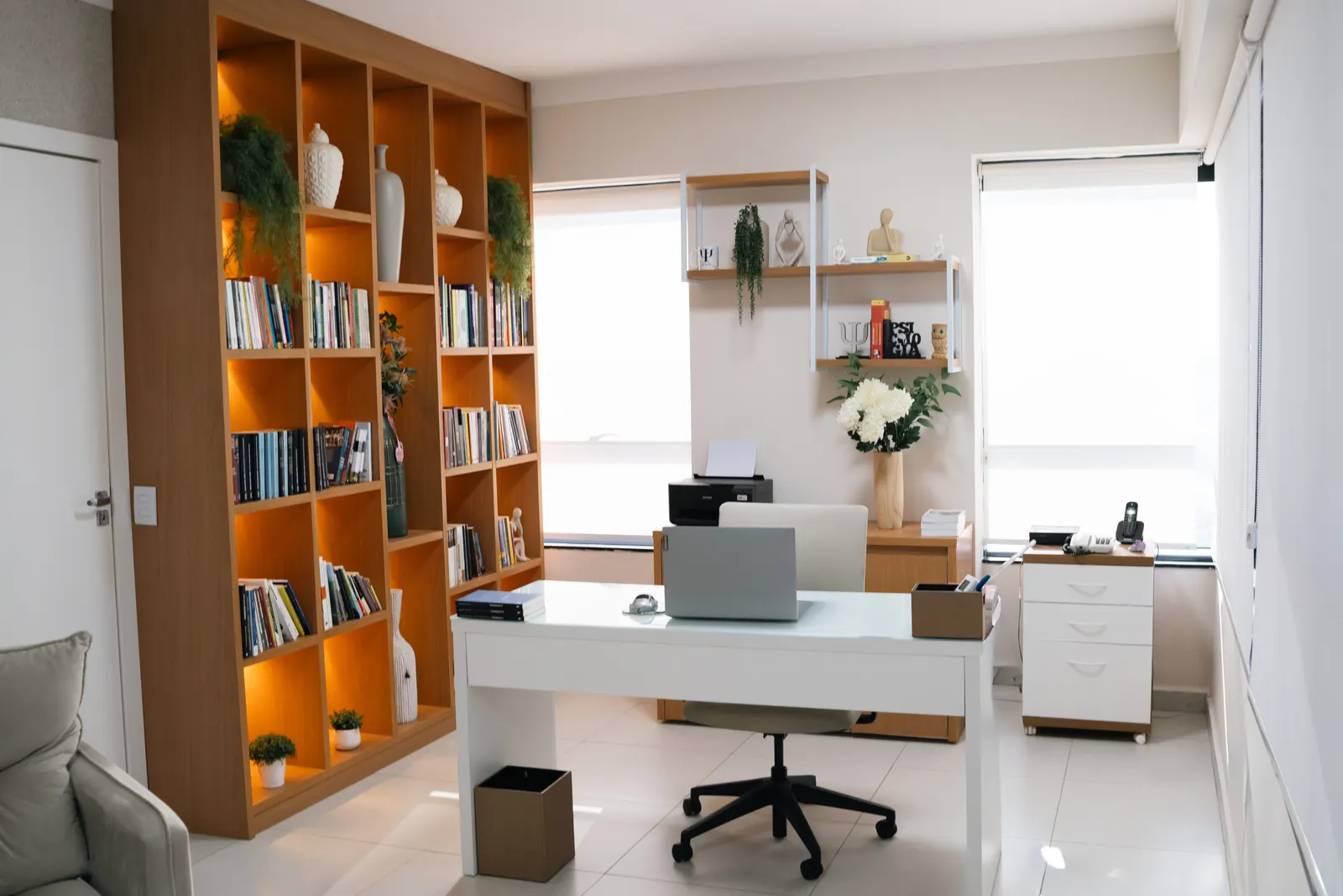 Sala com uma grande estante de madeira cheia de livros, vasos e plantas, mesa branca com computador e cadeira de escritório, iluminada por duas janelas amplas com persianas claras.